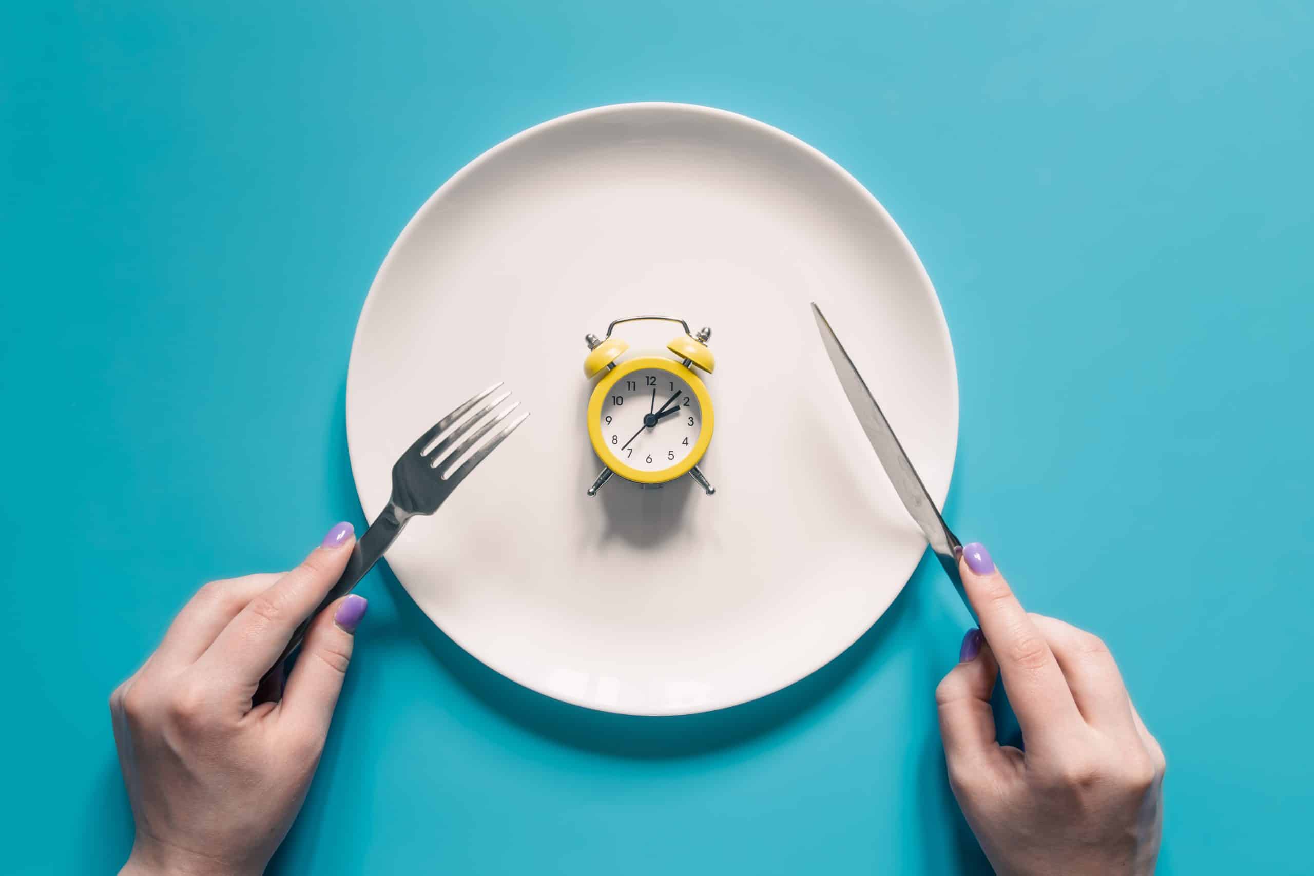 Hands holding knife and fork above alarm clock on a plate on blue background