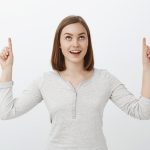 Waist-up shot of intrigued delighted good-looking european female in trendy clothes raising index fingers pointing up and smiling joyfully being interested with curious light in sky over gray wall